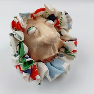 Könnte beinhalten: Ein skurriles Clown-Gesicht aus Keramik mit übertriebenen Merkmalen, darunter große, ausdrucksstarke Augen und eine markante Nase. Der Hals des Clowns ist mit einem Rüschenkragen aus Stoff mit roten, grünen, blauen und weißen Mustern verziert. Das Gesicht hat einen neutralen Hautton.