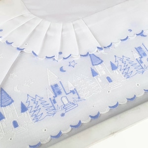 Puede incluir: Manta de bebé blanca con un diseño de castillo y árboles azules. La manta tiene un borde festoneado y un cuello con volantes. El diseño incluye castillos, árboles, estrellas y lunas bordados.