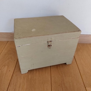 May include: A small, rectangular wooden chest painted in a muted green color. The chest has a hinged lid and a metal clasp. The wooden box sits on four small feet and is likely used for storage. The paint shows signs of wear, giving it a vintage appearance.