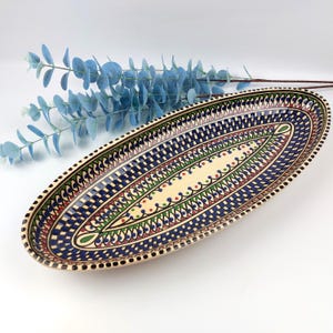 Op de afbeelding: Een ovale keramische serveerschaal met een crèmekleurig midden en een gedetailleerde rand. De rand heeft een blauw-wit geblokt patroon, met rode, groene en zwarte accenten. De schaal is ongeveer 40 cm lang.