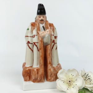 Puede incluir: Figura de cerámica de un hombre con un abrigo largo y un sombrero negro. El abrigo es de color crema con detalles rojos y verdes y ribetes marrones. La figura está sobre una base blanca.