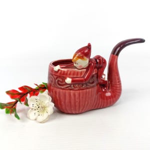 Op de afbeelding: Een rode keramische pijpvormige plantenbak met een kleine elfenfiguur die aan de zijkant klimt. De plantenbak heeft een textuuroppervlak en een gebogen steel. Een witte bloem en rode bladeren staan op de voorgrond. De elf draagt een rode hoed en outfit.