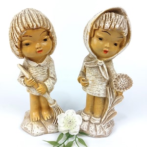 Puede incluir: Dos figuras de cerámica vintage de niños. Uno sostiene una escoba, el otro una flor. Ambos visten ropa texturizada de color blanquecino con piel bronceada y labios rojos. Las figuras se alzan sobre bases texturizadas, con una flor blanca delante.