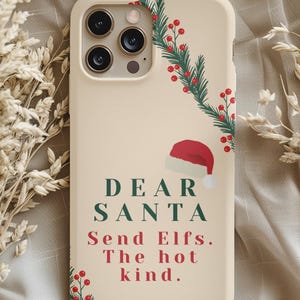 Könnte beinhalten: Beige Handyhülle mit Weihnachtsmotiv. Sie zeigt einen Weihnachtsmannhut, grüne Zweige mit roten Beeren und den Text "DEAR SANTA Send Elfs. The hot kind." Die Hülle hat einen Kameraausschnitt.