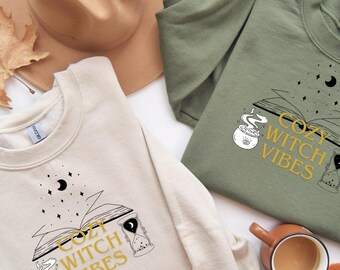 Könnte beinhalten: Zwei Sweatshirts, eines in Beige und eines in Olivgrün, mit dem Aufdruck "Cozy Witch Vibes" auf der Vorderseite. Das Design umfasst ein offenes Buch, einen Kessel, eine Sanduhr, Sterne und einen Mond. Ein brauner Hut und eine Kaffeetasse sind ebenfalls zu sehen.