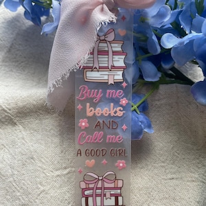 May include: Clear bookmark with pink text and illustrations of books and bows. The text reads "Buy me books AND Call me A GOOD GIRL". A pink ribbon is attached to the top of the bookmark. The bookmark is surrounded by blue and pink flowers.
