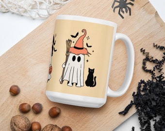 May include: A white ceramic mug featuring a graphic illustration of a ghost wearing an orange witch hat, holding a broomstick, and standing next to a black cat. The design includes small black bats and stars on a light beige background.