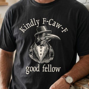 May include: Black t-shirt with a graphic of a crow wearing a top hat and tuxedo. The text above the crow reads "Kindly F-Caw-F", and below it reads "good fellow". The design is in a vintage style.
