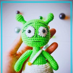 May include: A handmade green crochet alien doll with white eyes and a white skirt. The doll has small antennae and is held in a hand. The doll has a white strap with a yellow button.
