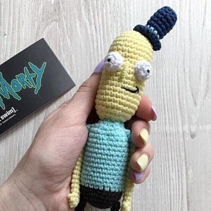 May include: A crocheted plush toy of a character from the animated series Rick and Morty. The toy is yellow with a blue hat, blue shirt, black pants, and brown shoes. The toy has a smiling face with large white eyes.