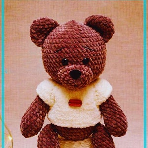 May include: A brown crocheted teddy bear wearing a white crocheted shirt with a red button. The bear is sitting on a white wicker stool.