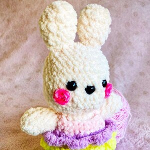 Könnte beinhalten: Ein gehäkeltes Hasenspielzeug mit rosa Wangen und schwarzen Augen. Der Hase trägt einen mehrfarbigen Rock mit rosa, lila, gelben und blauen Schichten.