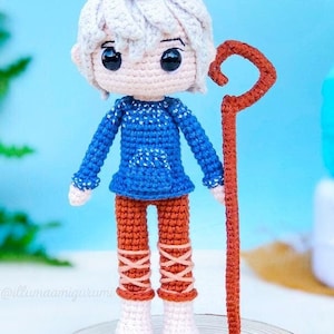 Peut inclure: Une poupée amigurumi au crochet de Jack Frost du film Le  Gardien des rêves. La poupée porte un pull bleu avec des flocons de neige blancs, un pantalon marron et des bottes blanches. La poupée tient un bâton brun avec un sommet incurvé.