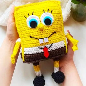 Könnte beinhalten: Ein gehäkeltes SpongeBob Schwammkopf Spielzeug mit einem gelben Körper, braunen Hosen, einem weißen Hemd und einer roten Krawatte. Das Spielzeug hat ein lächelndes Gesicht mit blauen Augen und weißen Pupillen.