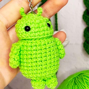 Könnte beinhalten: Ein hellgrüner gehäkelter Schlüsselanhänger in Form eines Android-Roboters. Der Schlüsselanhänger hat schwarze Augen und eine silberne Kette.