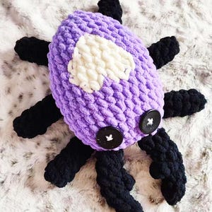 May include: A purple crocheted spider with black legs and white accents. The spider has two black buttons for eyes.