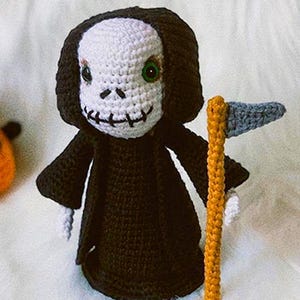 Puede incluir: Un juguete de peluche tejido a crochet de la Muerte, con una túnica negra con capucha y sosteniendo una guadaña. El juguete tiene una cara blanca con ojos verdes y una sonrisa cosida.