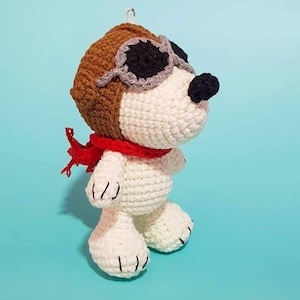Puede incluir: Un perro de crochet blanco que lleva gafas de aviador marrones y una bufanda roja. El perro tiene ojos negros y una nariz negra.