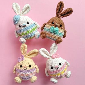 Könnte beinhalten: Vier gehäkelte Osterhasen-Plüschtiere, jedes in Form eines Eies und mit bunten Streifen verziert. Die Hasen sind weiß, braun, gelb und weiß mit rosa Akzenten.