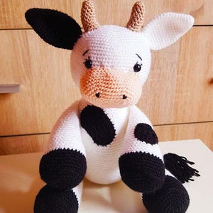 May include: A crocheted stuffed cow toy with black and white spots. The cow has a white body, black hooves, and a black tail. The cow's eyes are black with white pupils. The cow's nose is pink.