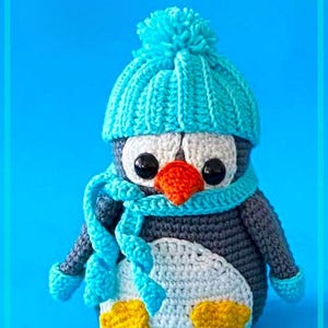 May include: A crocheted penguin wearing a blue hat and scarf. The penguin has black eyes, an orange beak, and white and gray body with yellow feet.