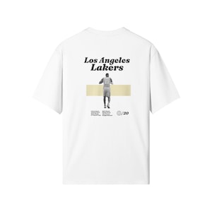 May include: White t-shirt featuring the text "Los Angeles Lakers" in a vintage font. The back of the shirt displays a black and white image of a basketball player. Additional text and a globe icon are also present.