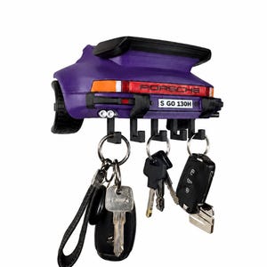 May include: A purple wall-mounted key holder shaped like the rear of a Porsche sports car. It features a black spoiler, orange and red taillights, and a license plate reading S GO 130H. Five black hooks hold several metal keys and key fobs.