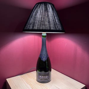 May include: A unique table lamp crafted from a champagne bottle, featuring a black lampshade. The bottle has a dark green hue with a label that reads "KRUG". The lamp is placed on a wooden surface against a burgundy wall.