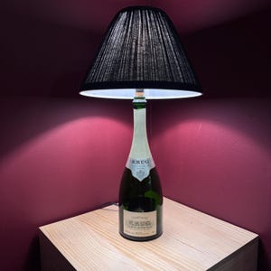 May include: A unique table lamp made from a champagne bottle, featuring a black pleated lampshade. The bottle is green with a white label that reads "KRUG CHAMPAGNE". The lamp sits on a light-colored wooden surface.