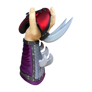 Puede incluir: Un mando de videojuegos rojo y negro sostenido por una escultura con forma de mano con garras plateadas y un brazalete morado y plateado. La escultura tiene un diseño de cota de malla.