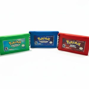 Może przedstawiać: Trzy kolorowe breloczki do gier Pokémon: zielony "Emerald Version", niebieski "Sapphire Version" i czerwony "Ruby Version". Każdy breloczek ma tytuł gry i czarny łańcuszek kulkowy. Kartridże są prostokątne i mają błyszczące wykończenie.