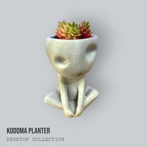 May include: A light gray Kodama planter with a seated figure design, holding its knees. The head of the figure is a planter filled with a succulent plant with green, yellow, and red leaves. The text "KODAMA PLANTER DESKTOP COLLECTION" is at the bottom.