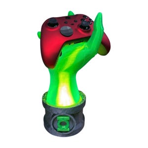 Op de afbeelding: Een groene en zwarte gamecontrollerstandaard in de vorm van een hand die een rode controller vasthoudt. De basis heeft een groen logo. De standaard is ontworpen om een gamecontroller vast te houden.