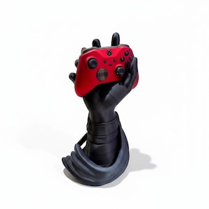 May include: A red Xbox controller held by a black, stylized hand-shaped stand. The stand has a textured, armored design. The controller features black buttons and a checkered pattern on the center button, set against a white background.