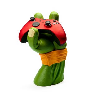 May include: A green, stylized hand-shaped gaming controller stand with an orange band around the wrist. A red gaming controller rests in the hand. The stand is designed to hold a gaming controller.
