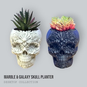 May include: Two skull-shaped planters, one white and one dark blue, each holding a succulent plant. The planters are decorated with floral patterns. The text "MARBLE & GALAXY SKULL PLANTER" and "DESKTOP COLLECTION" is at the bottom.