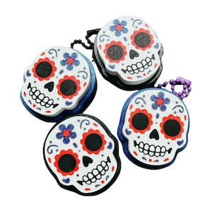 May include: Four Day of the Dead skull-shaped coin purses. Each purse features a white skull design with red, blue, and black accents. The purses have a blue glittery border and a zipper closure. Some have a chain.
