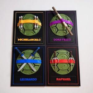 Puede incluir: Cuatro piezas de arte mural enmarcadas con diseños de las Tortugas Ninja. Cada pieza tiene un caparazón de tortuga verde con una máscara y un arma de color. Los nombres Michelangelo, Donatello, Leonardo y Raphael se muestran debajo.