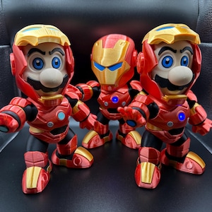 May include: Three figurines of Mario characters in Iron Man armor. The figures are red, gold, and black, with blue eyes and glowing chest pieces. The helmets have gold accents.