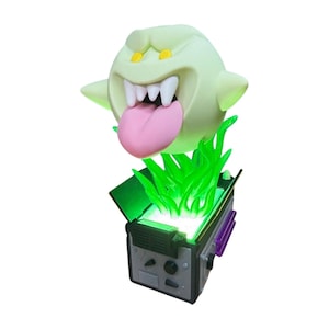 May include: A light green cartoon ghost figure with a pink tongue and sharp teeth, above a grey and black box emitting bright green light. The box has purple accents and a control panel.