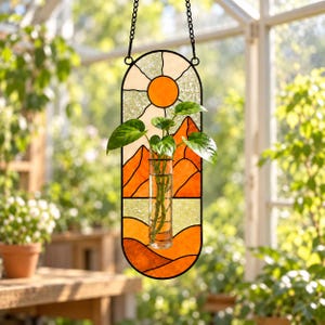 Könnte beinhalten: Ein Wandbehang aus Buntglas mit einer orangefarbenen Sonne und einer Berglandschaft, in dessen Mitte eine Reagenzglasvase aus Klarglas befestigt ist. Das vertikale ovale Paneel hängt an einer schwarzen Metallkette.