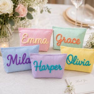 May include: Five rectangular zipped pouches made of pinstriped fabric in pink, light green, light blue, lavender, and yellow. Each pouch features a name embroidered in a contrasting colour: Emma in yellow, Grace in orange, Mila in purple, Harper in teal, and Olivia in teal.