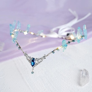 May include: A silver headband with a blue crystal pendant and light blue crystal accents. The headband is tied with a white ribbon.
