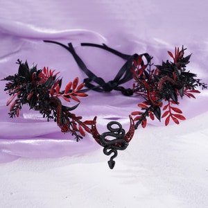 May include: A black and red floral crown with a crescent moon and snake design. The crown is made of plastic and has a black ribbon tie.
