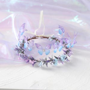 May include: A delicate floral crown featuring lavender and blue flowers, with several translucent butterfly accents. The crown is adorned with small, light blue flowers and purple berries, and has a white ribbon tie.