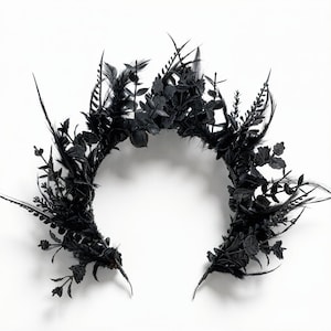 May include: A black floral crown featuring feathers and botanical elements. The crown is adorned with dark leaves, flowers, and feathers, creating a gothic or fantasy aesthetic. The design is symmetrical and set against a white backdrop.