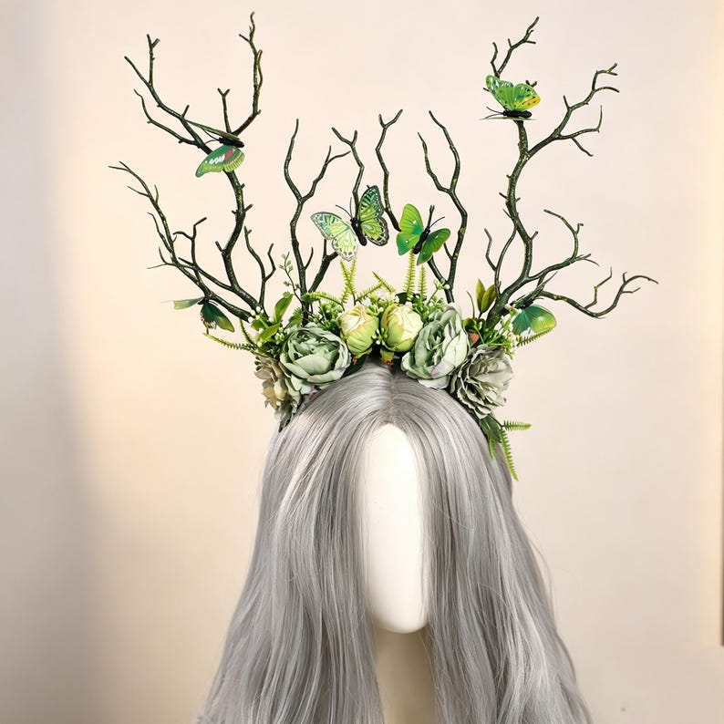 May include: A decorative headpiece featuring dark green branches, green and white flowers, and green butterfly accents. The headpiece is designed to sit atop a mannequin head with long, gray hair. The overall theme is nature-inspired.