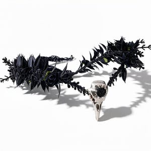 Op de afbeelding: Een zwarte bloemenkrans in gotische stijl met een zilveren vogel schedel hanger. De kroon heeft donkere bloemen en bladeren, met een gedetailleerde schedel hanger aan de voorkant. Het ontwerp staat tegen een witte achtergrond.