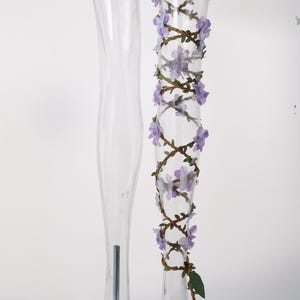 May include: A clear, transparent mannequin leg display stand featuring a decorative brown vine wrap adorned with small, light purple fabric flowers and green leaves that spiral around the length of the leg from top to bottom.