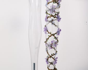 Könnte beinhalten: Ein transparenter Schaufensterpuppen-Beinständer, der mit einer dekorativen braunen Ranke umwickelt ist, die mit kleinen hellvioletten Stoffblumen und grünen Blättern verziert ist und sich von oben nach unten um das Bein windet.